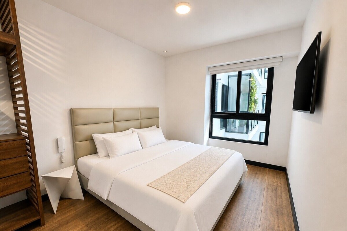 A hotel room with a bed, a bedside table, a wall-mounted TV, and a window with a view of buildings.
