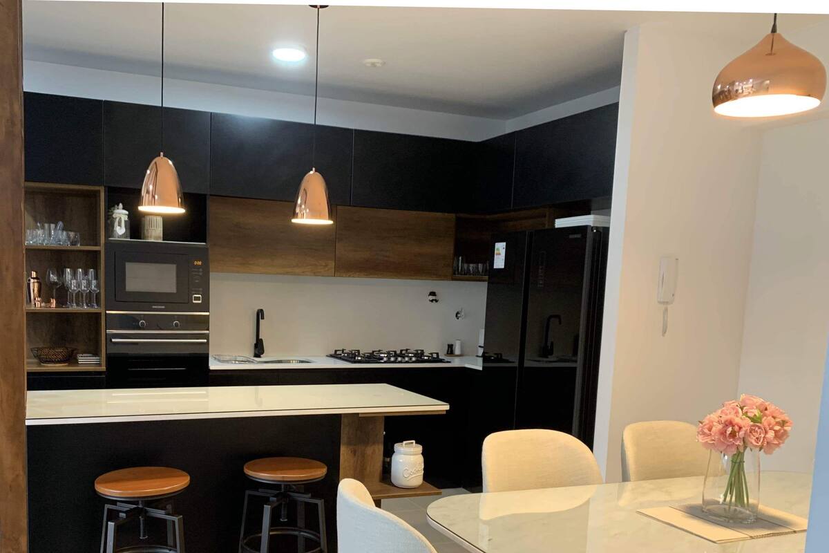 Een moderne keuken met zwarte kastjes, een wit aanrechtblad en een centraal eiland met krukken. Boven het eiland hangen hanglampen en op de eettafel staat een vaas met bloemen.