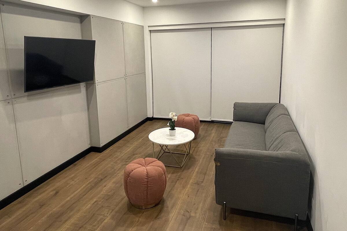 Een moderne woonkamer met een grijze bank, een ronde witte salontafel en twee roze poefs. Een flatscreen televisie is aan de muur bevestigd, en er is een schuifdeur in de achtergrond te zien.