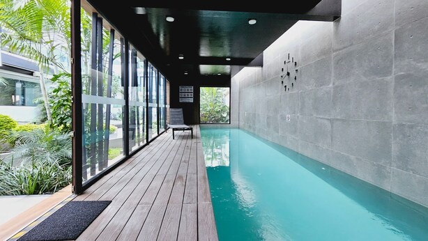 A modern indoor pool area with a wooden deck, large windows, and a view of greenery outside.