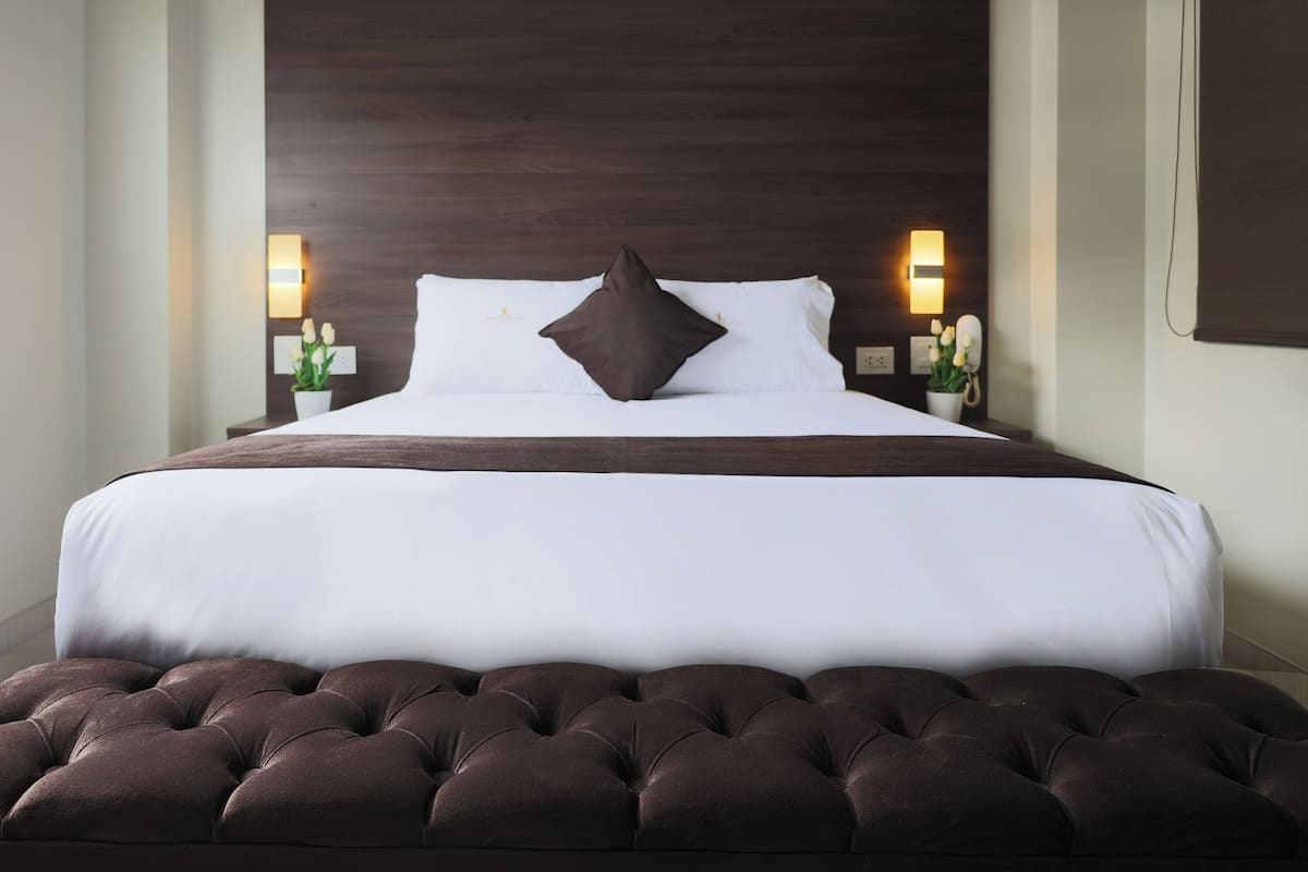 A neatly made bed with a dark brown headboard, white linens, and a single dark brown pillow. There are two bedside tables with lamps and small potted plants.