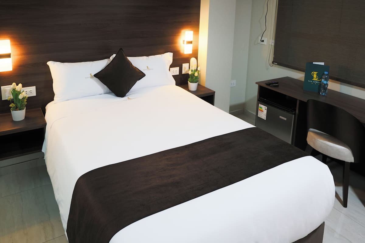 A hotel room with a large bed, two bedside tables with lamps, a desk with a chair, and a TV on the wall.