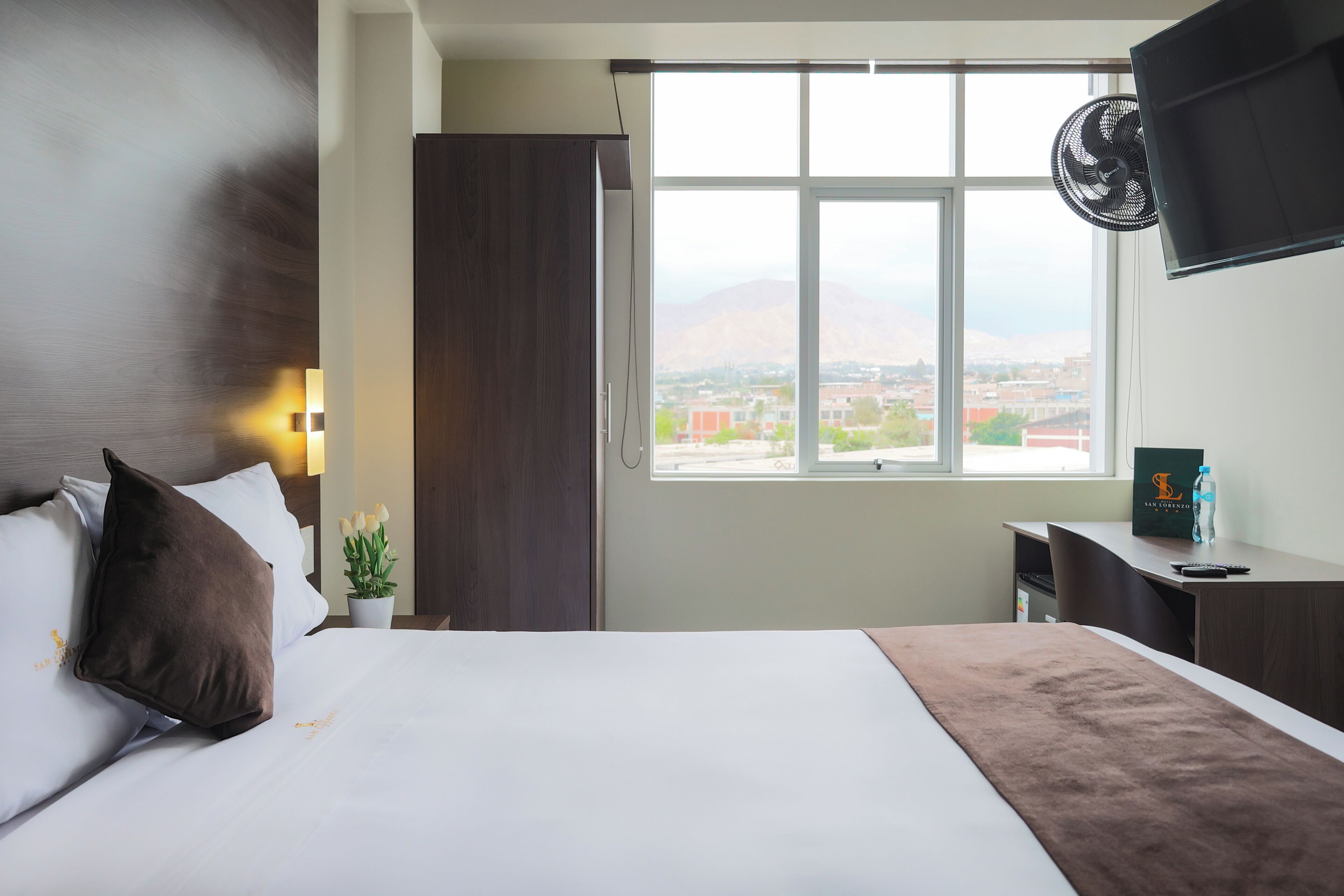 Habitación de hotel con cama, escritorio, televisión, ventana con vista a edificios y montañas, y un ventilador.