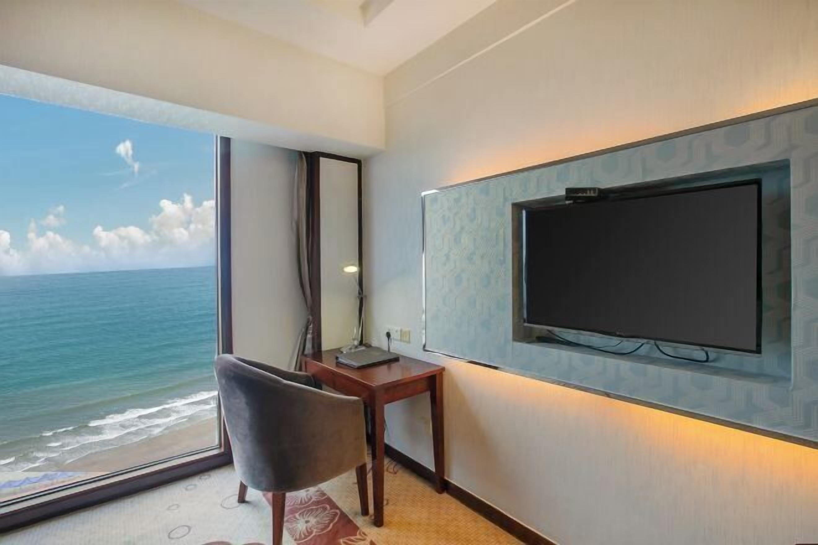 Quarto de hotel com uma janela ampla vista para o mar, televisão de tela plana na parede, escrivaninha de madeira e uma cadeira.