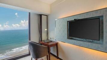 Quarto de hotel com uma janela ampla vista para o mar, televisão de tela plana na parede, escrivaninha de madeira e uma cadeira.