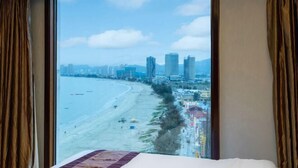 A hotel room with a large window offering a view of a beach and cityscape. - Haibaina Hotel (Huidong)