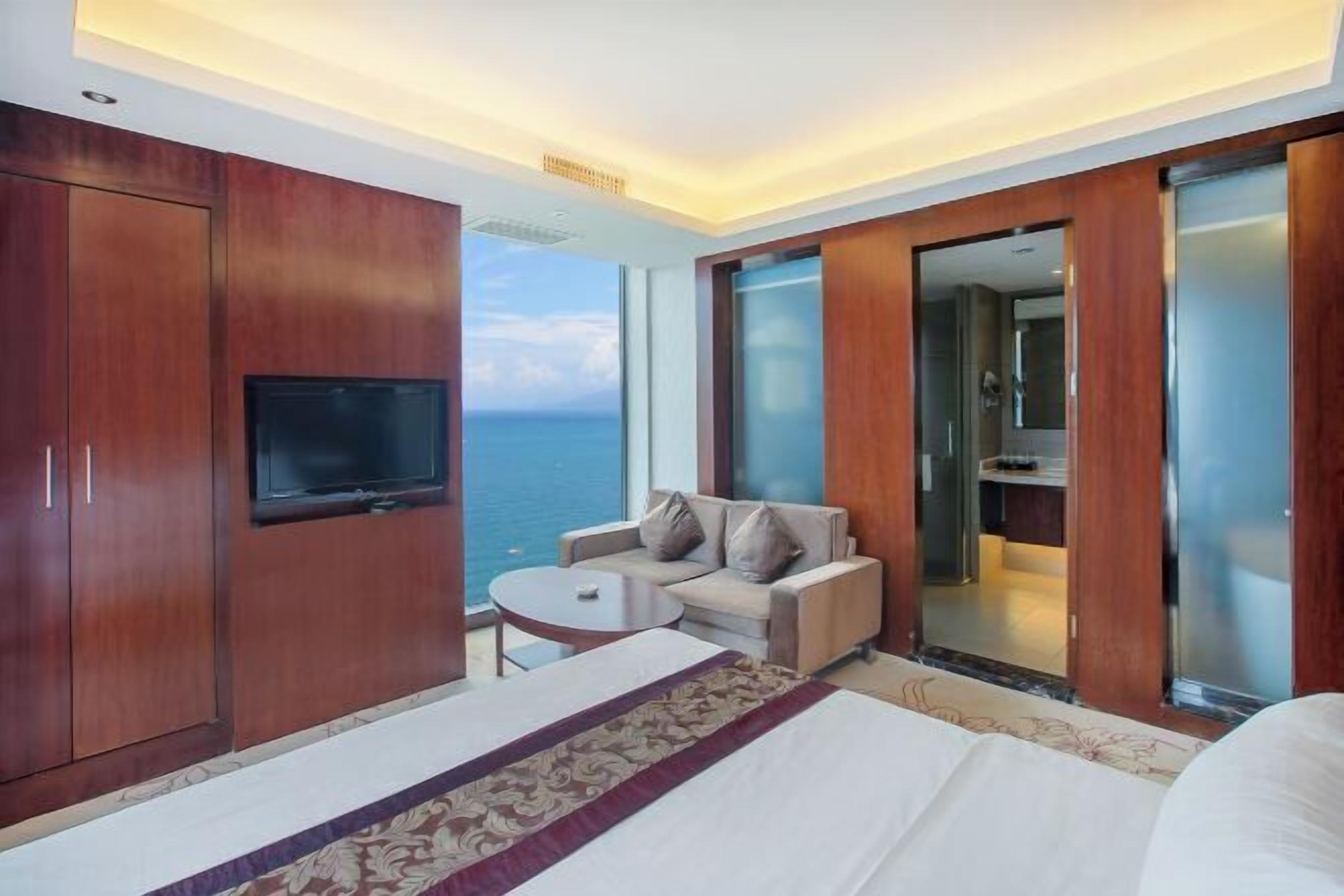 A hotel room with a bed, a TV, a sofa, a small table, and a view of the sea.