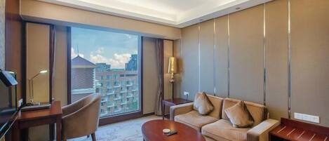 A hotel room with a sofa, a coffee table, a desk, and a large window offering a city view.