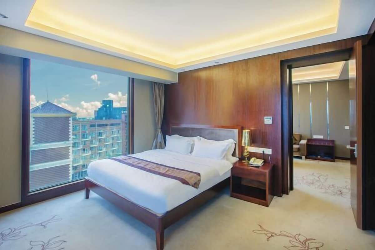 A hotel room with a large bed, a nightstand, a lamp, and a view of a cityscape through a large window.