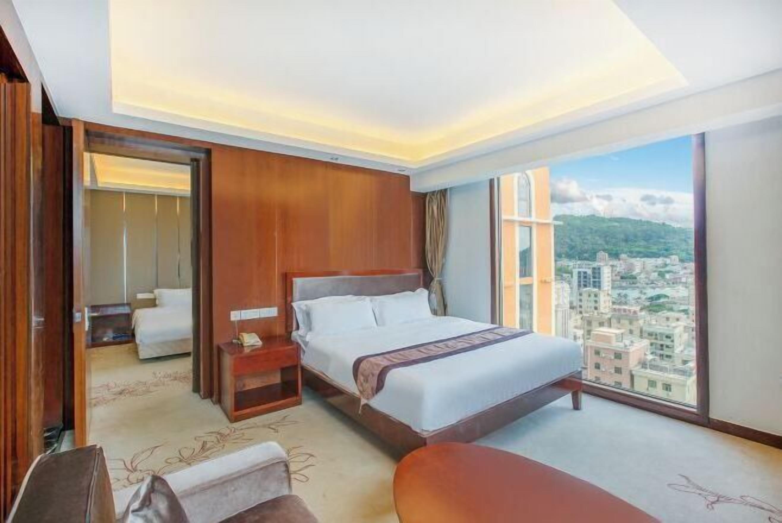Quarto de hotel com uma cama grande, uma mesinha de cabeceira, uma cadeira e vista para a cidade através de amplas janelas.