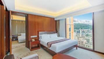 Quarto de hotel com uma cama grande, uma mesinha de cabeceira, uma cadeira e vista para a cidade através de amplas janelas.