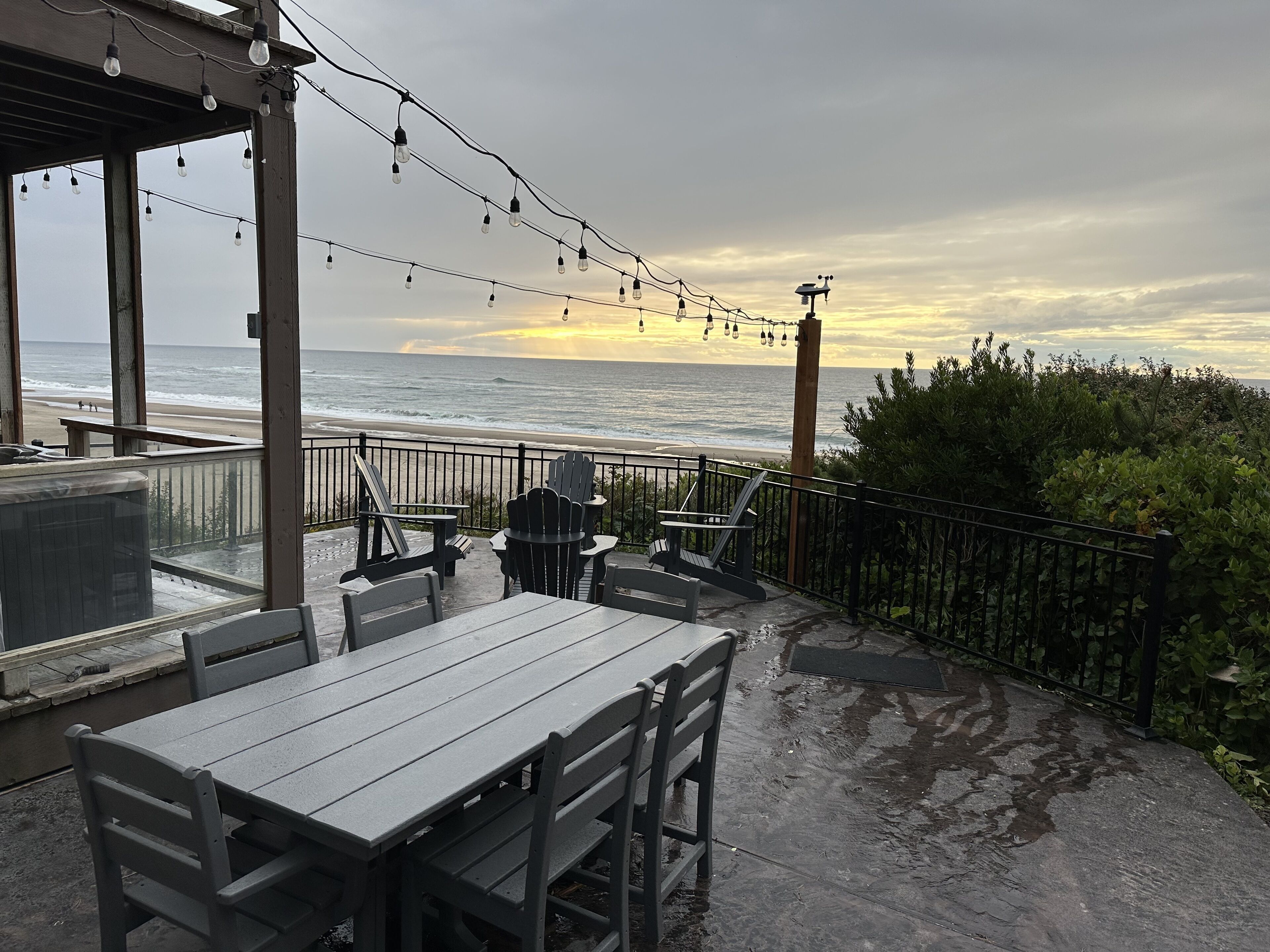 An outdoor patio with a view of the ocean, featuring a wooden table and chairs, string lights, and a railing.