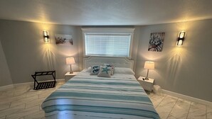 A bedroom with a large bed, two bedside tables with lamps, a bench, a window with blinds, and wall-mounted artwork. - Luxury beachfront property with amazing views,  hot tub and steps to the beach.  (Lincoln Beach)