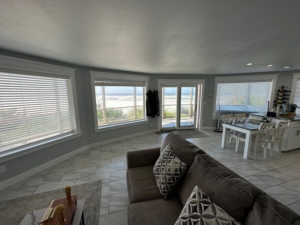 A spacious living room with a large gray sofa, a dining area with white chairs, and a kitchen with a countertop. - Luxury beachfront property with amazing views, hot tub and steps to the beach. (Lincoln Beach)