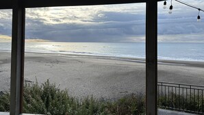 A beach view from a covered area with a railing, featuring a cloudy sky and calm sea. - Luxury beachfront property with amazing views,  hot tub and steps to the beach.  (Lincoln Beach)