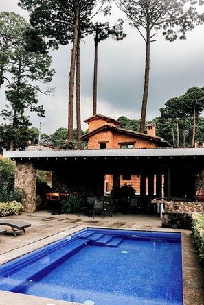 A swimming pool with a stone deck, surrounded by tall trees and a brick building.