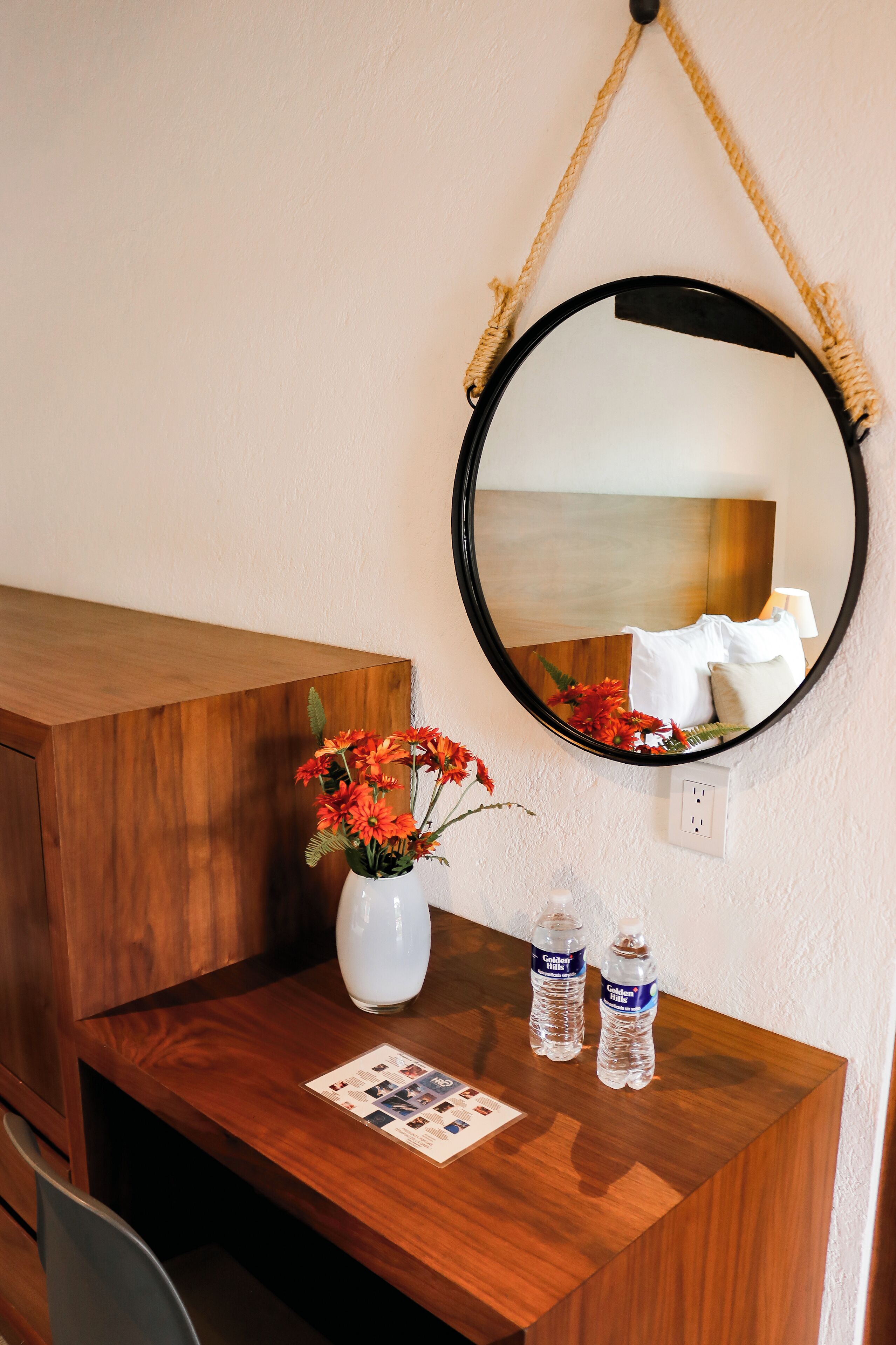 Un bureau en bois avec un miroir, un vase rempli de fleurs et deux bouteilles d’eau.