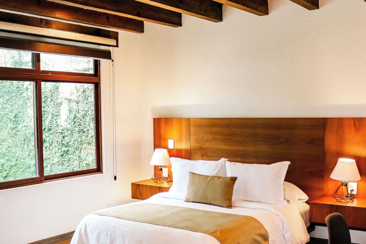 Un dormitorio con techo de madera, una cama grande, dos lámparas de noche y vistas al verdor a través de una ventana.