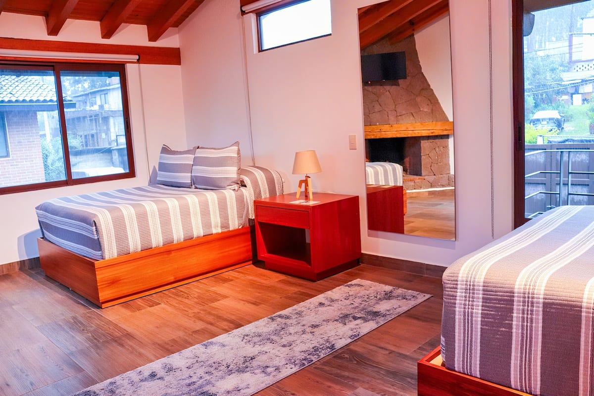 Un dormitorio con una cama de madera, una mesita de noche roja, una lámpara, un ventanal grande y vistas a una chimenea de piedra.