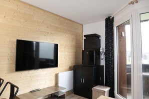 Un salon moderne avec un téléviseur à écran plat fixé sur un mur en bois, un meuble noir, une petite table et une porte-fenêtre coulissante donnant sur un espace extérieur.