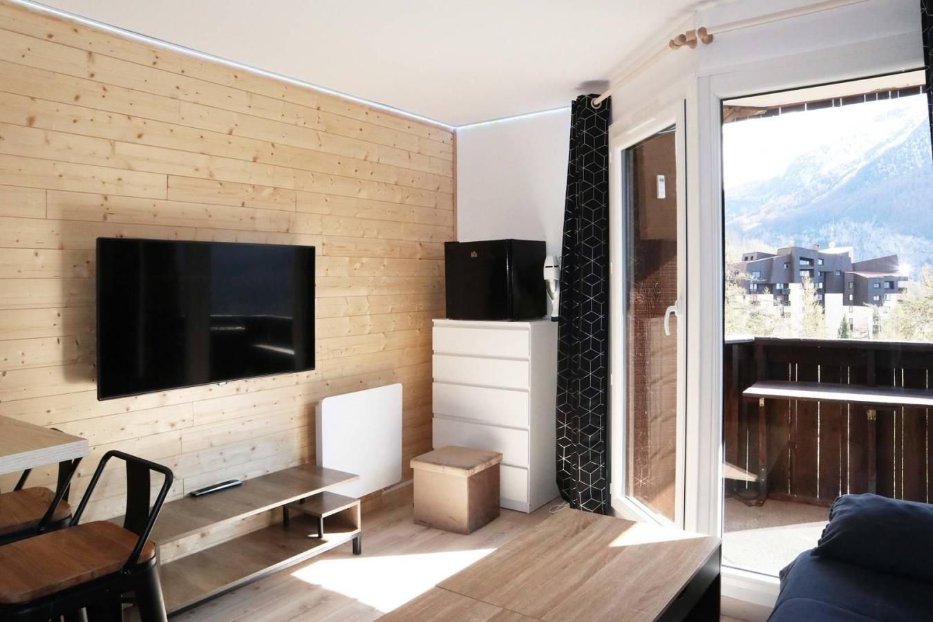Ein modernes Wohnzimmer mit Fernseher, eine Essecke mit Tisch und Stühlen sowie ein Balkon mit Blick auf Gebäude und Berge.