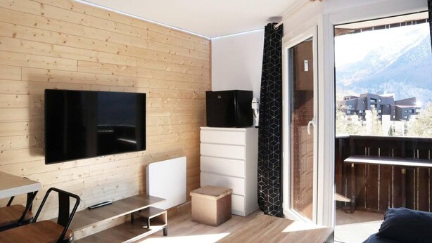En moderne stue med et fladskærms TV monteret på en trævæg, et spiseområde med bord og stole, et lille hvidt skab og en balkon med udsigt til bygninger og bjerge.