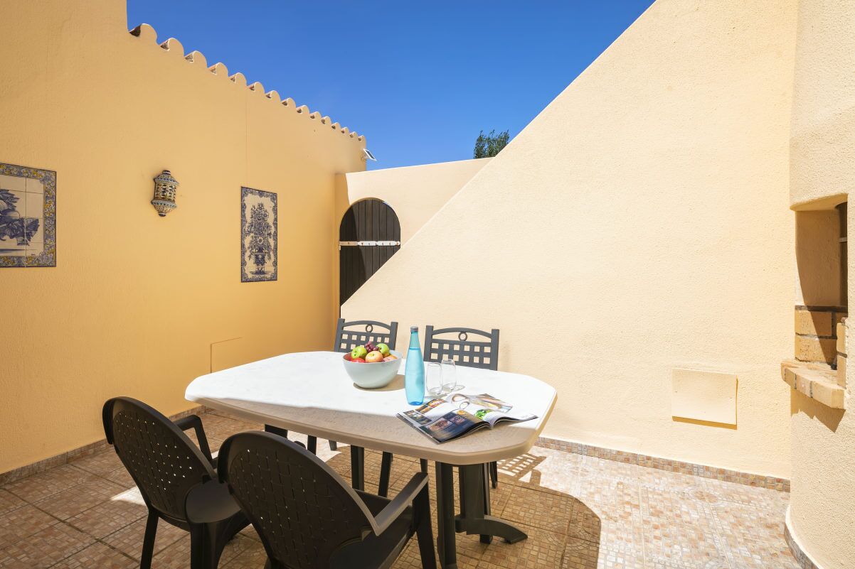 A small outdoor dining area with a table and chairs, a bowl of fruit, a water bottle, and a magazine.