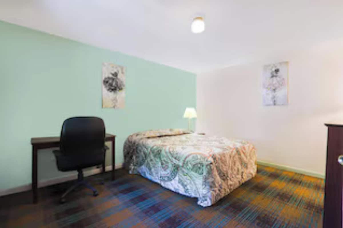 A room with a bed, a desk, and a chair. There are two wall-mounted artworks.