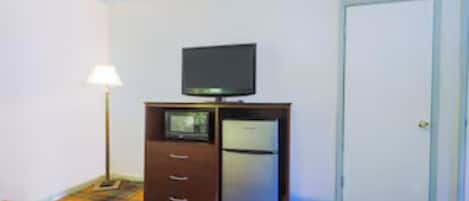 Habitación de hotel con cama, televisor sobre un mueble de madera, refrigerador, lámpara y puerta.