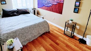 Ein Schlafzimmer mit einem Bett, einem Fenster, einem an der Wand befestigten Luftreiniger, einem kleinen Tisch mit einer Pflanze und einer Wand mit gerahmten Bildern.