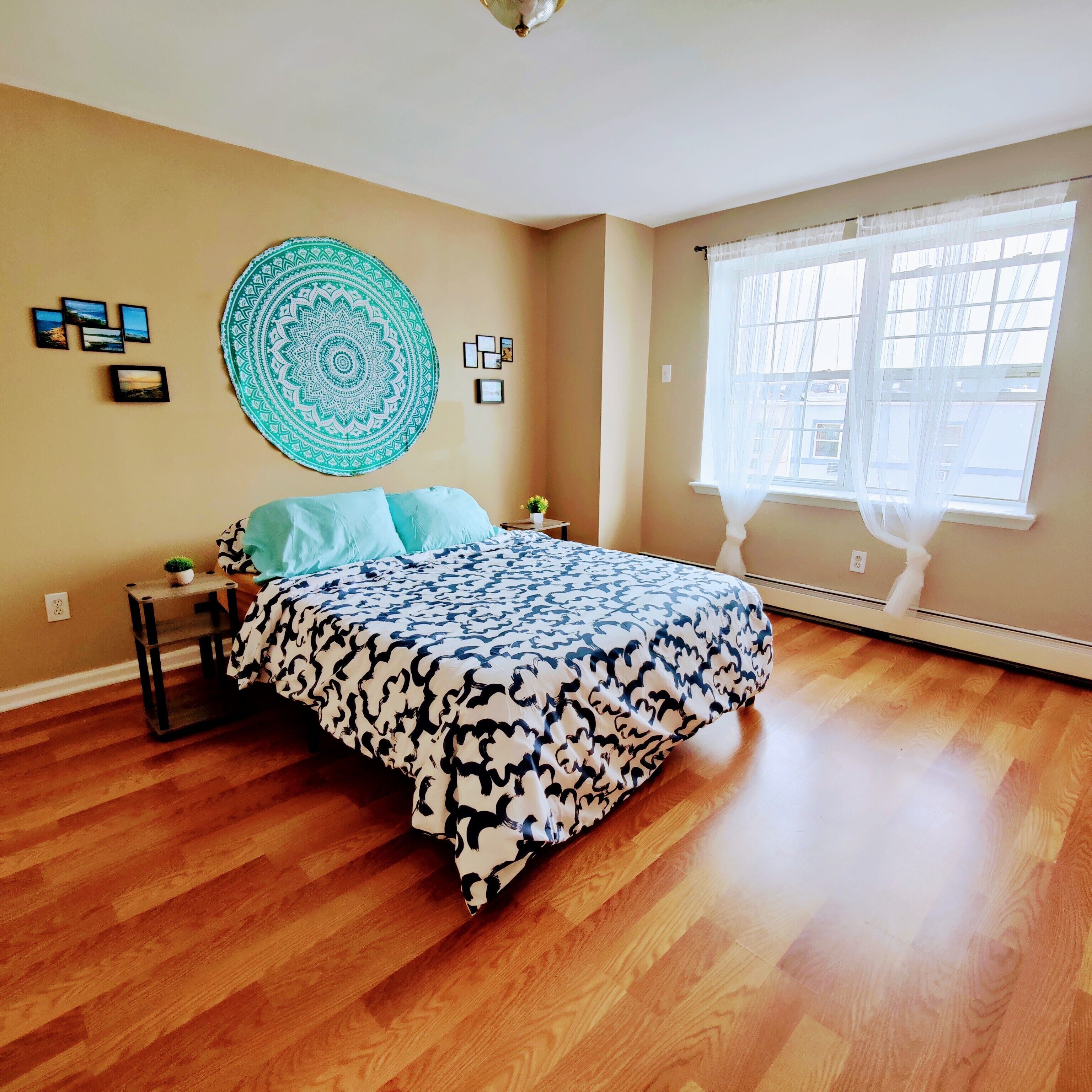 A bedroom with a bed, a round wall tapestry, framed pictures, and a window with sheer curtains.