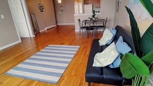 Ein geräumiges Wohnzimmer mit Holzboden, einem blau gestreiften Teppich, einem dunklen Sofa mit gemusterten Kissen und einer großen Grünpflanze.