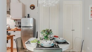 A dining area with a round table set for four, surrounded by chairs. A refrigerator and kitchen area are visible in the background.