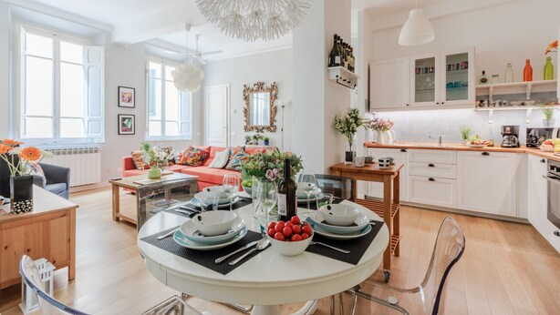 A modern living room with a round dining table set for four, a kitchen with white cabinets, and a large window providing natural light.