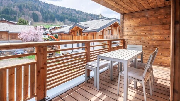 Eine Holzterrasse mit Tisch und Stühlen mit Blick auf eine gebirgige Landschaft und einige Häuser.