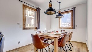 A dining area with a wooden table set for four, surrounded by brown chairs. There are two windows with curtains, and a ceiling-mounted light fixture above the table. - 140m from ski lifts, 1st floor, terrace, tv, 77m², Morzine (Morzine)