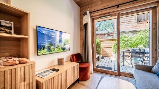 A cozy room with a wooden ceiling, a large window with a view of a wooden deck and greenery, a flat-screen TV mounted on the wall, and a wooden reception desk with a magazine and a glass of drink.