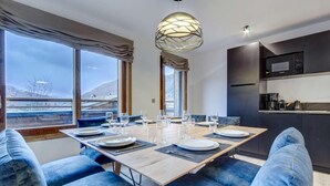 A modern dining area with a wooden table set for four, blue upholstered chairs, and a large window offering a view of a snowy landscape.
