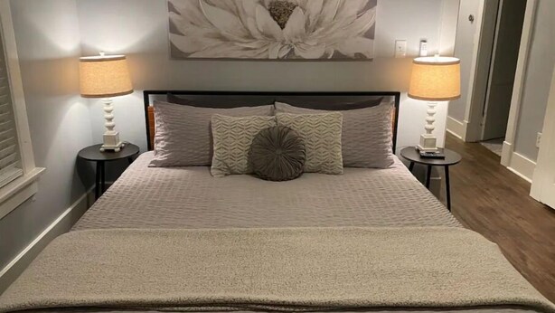 Ein ordentlich bezogenes Bett mit einem großen floralen Bild darüber, zwei Nachttischlampen und einer gemusterten Tagesdecke.
