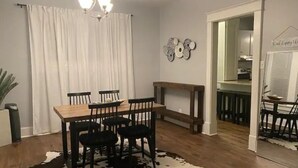 Ein Essbereich mit Holztisch und -stühlen, einem Kuhfellteppich, einer Deckenleuchte und einer durch einen offenen Durchgang sichtbaren Küche.
