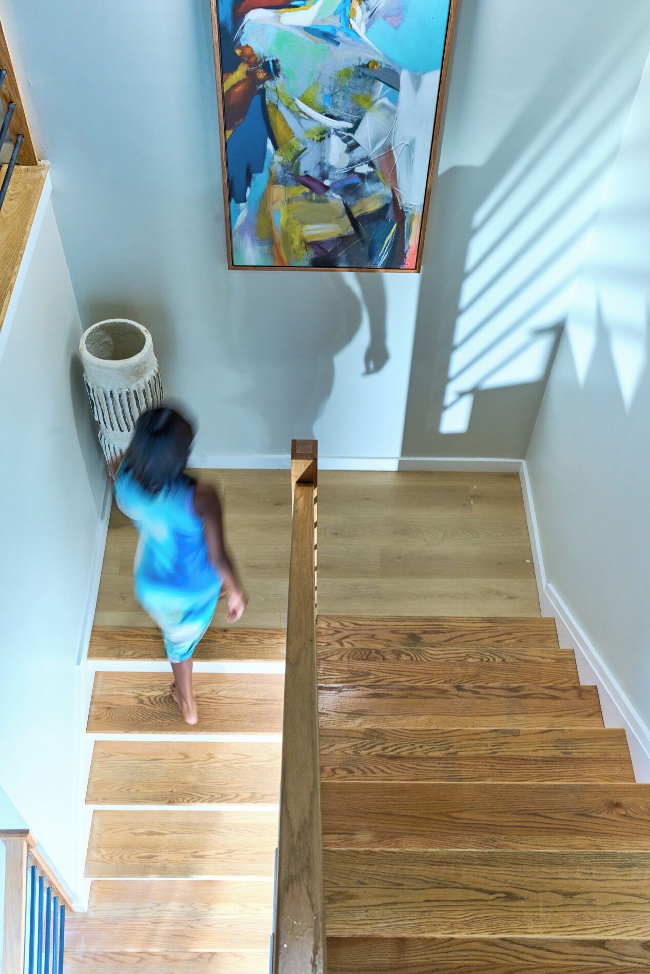 A wooden staircase with a handrail, a wall-mounted abstract painting, and a white trash bin.