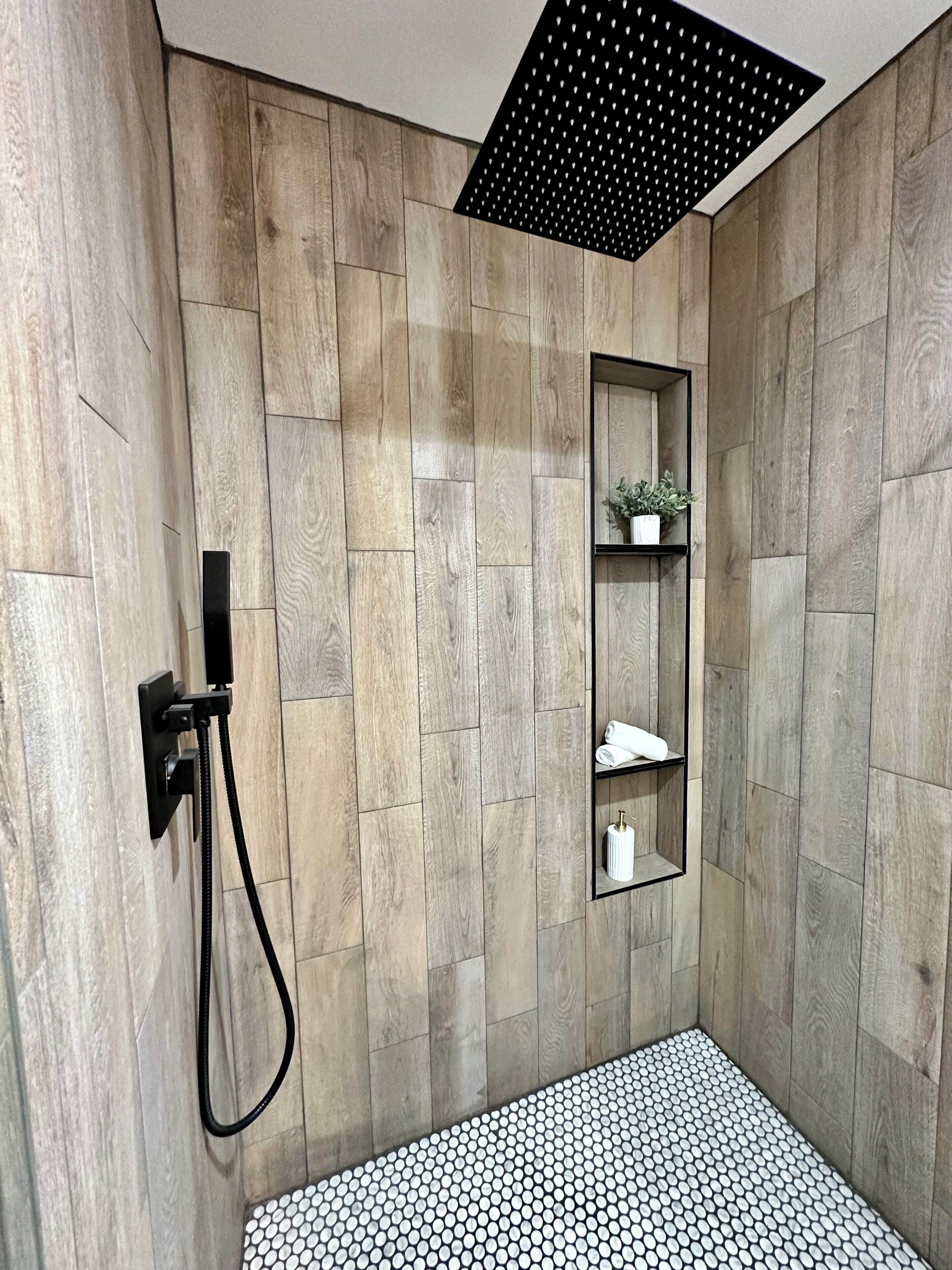 A shower with wooden paneling, a black ceiling vent, a shelf with a plant, and a tiled floor.