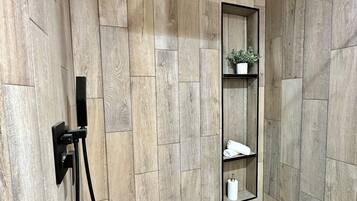 A shower with wooden paneling, a black ceiling vent, a shelf with a plant, and a tiled floor.