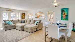 A spacious living room with a beige sectional sofa, a dining table with white chairs, and a ceiling fan.