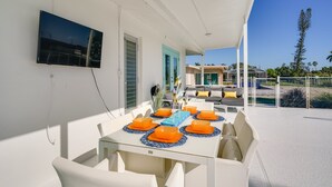A covered outdoor dining area with white tables and orange placemats, a TV mounted on the wall, and a view of a house and palm trees in the background.
