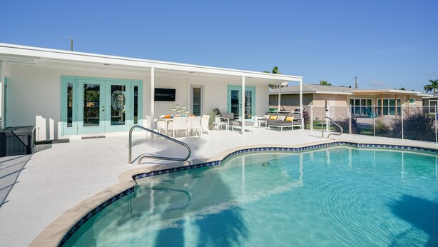 A swimming pool with a tiled edge, a covered patio area with outdoor furniture, and a building with teal-framed windows.