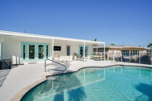 A swimming pool with a tiled edge, a covered patio area with outdoor furniture, and a building with teal-framed windows. - Dock, Grill & Pool: Waterfront Cape Coral Home! (Cape Coral)