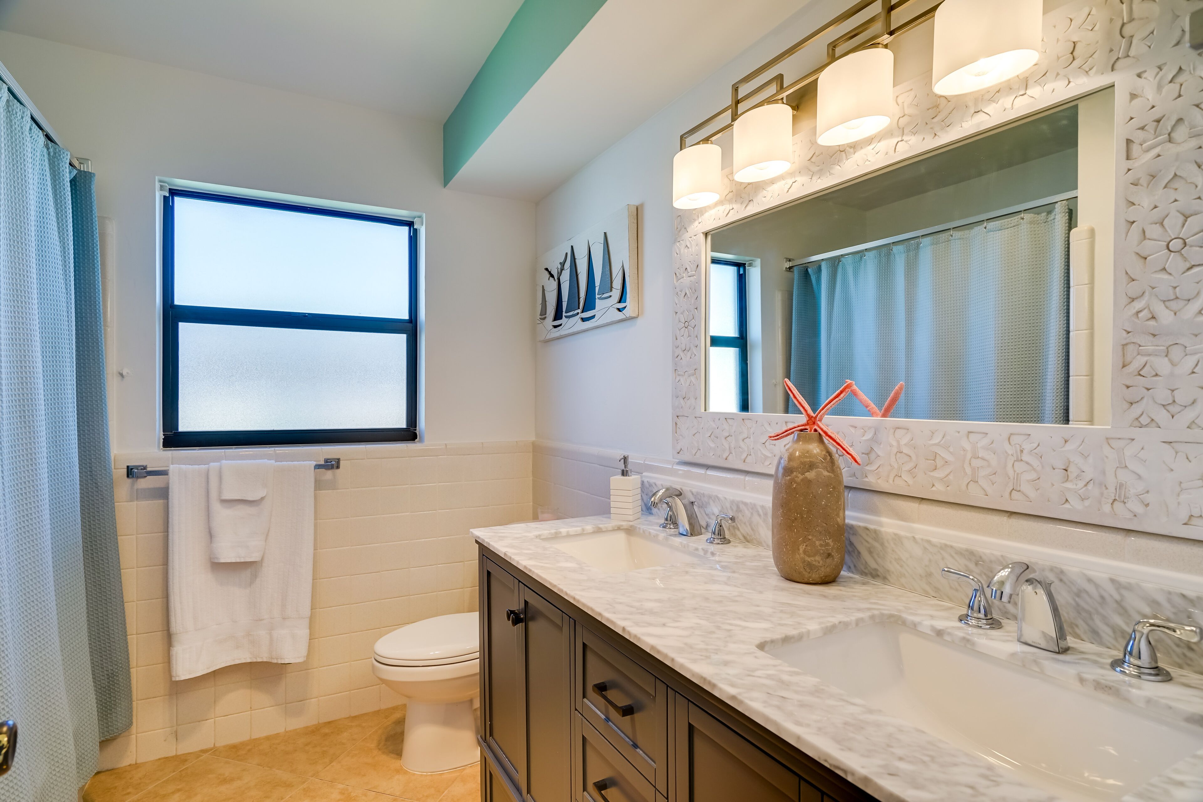 Un baño con inodoro blanco, lavamanos con doble grifo, un espejo grande con iluminación y una ventana con cortina.