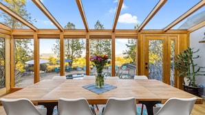 Un coin repas avec une table en bois et des chaises blanches, un vase de fleurs et une vue sur l’extérieur grâce à de grandes fenêtres.