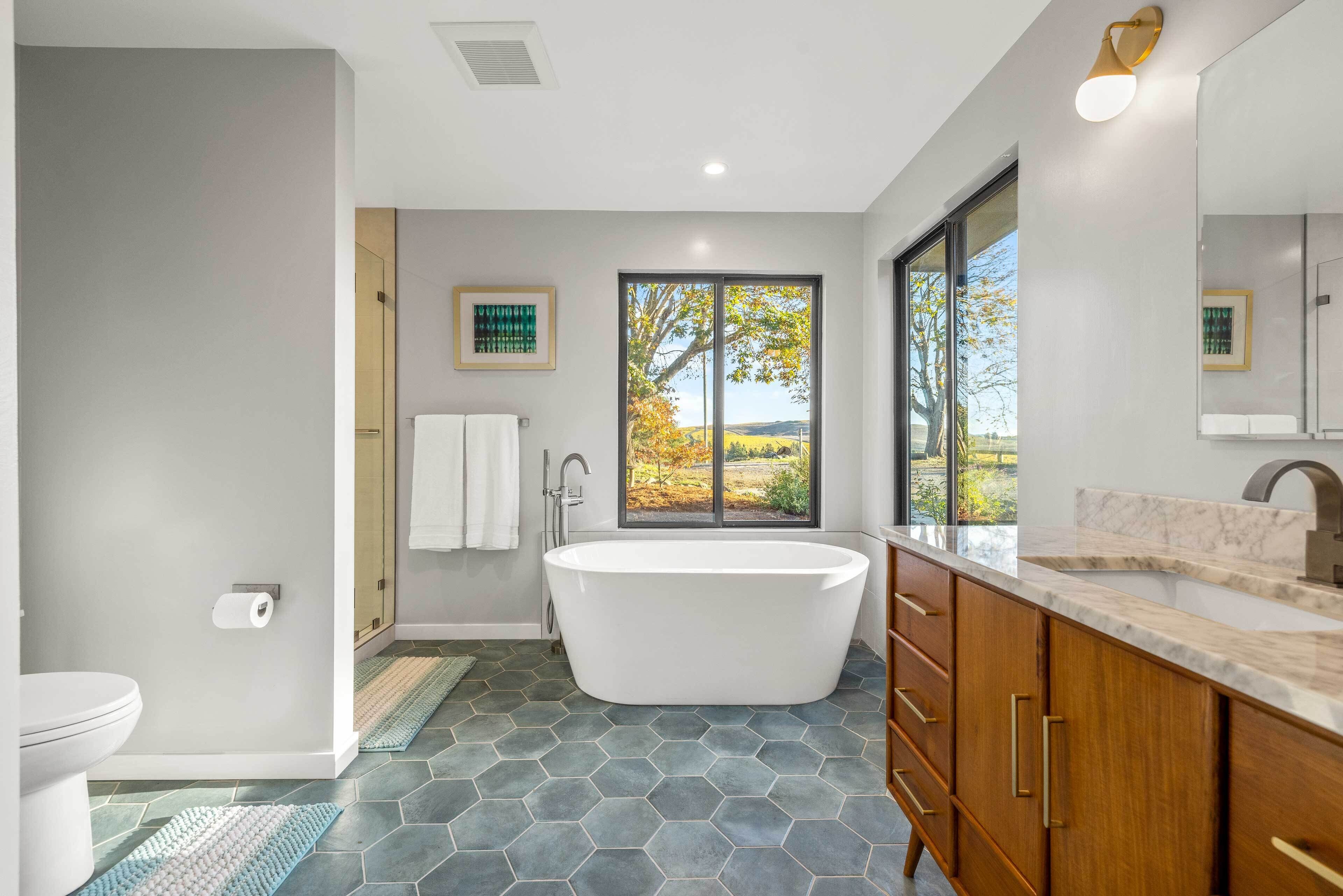 A modern bathroom with a freestanding bathtub, a large window with a view of trees, and a marble countertop.